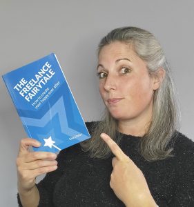 A photograph of Lisa Slater holding a copy of her book - The Freelance Fairytale - in one hand and pointing to it with the other.